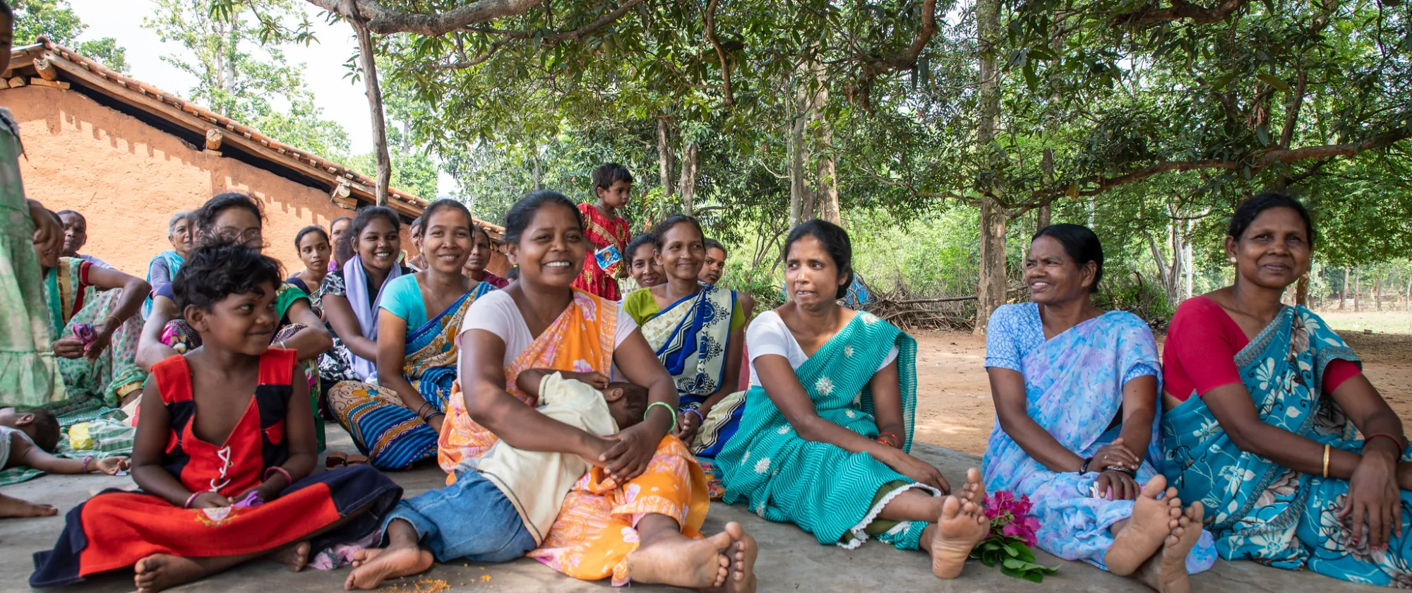 A group of women and children sit together outdoors under the shade of large trees in a rural village.