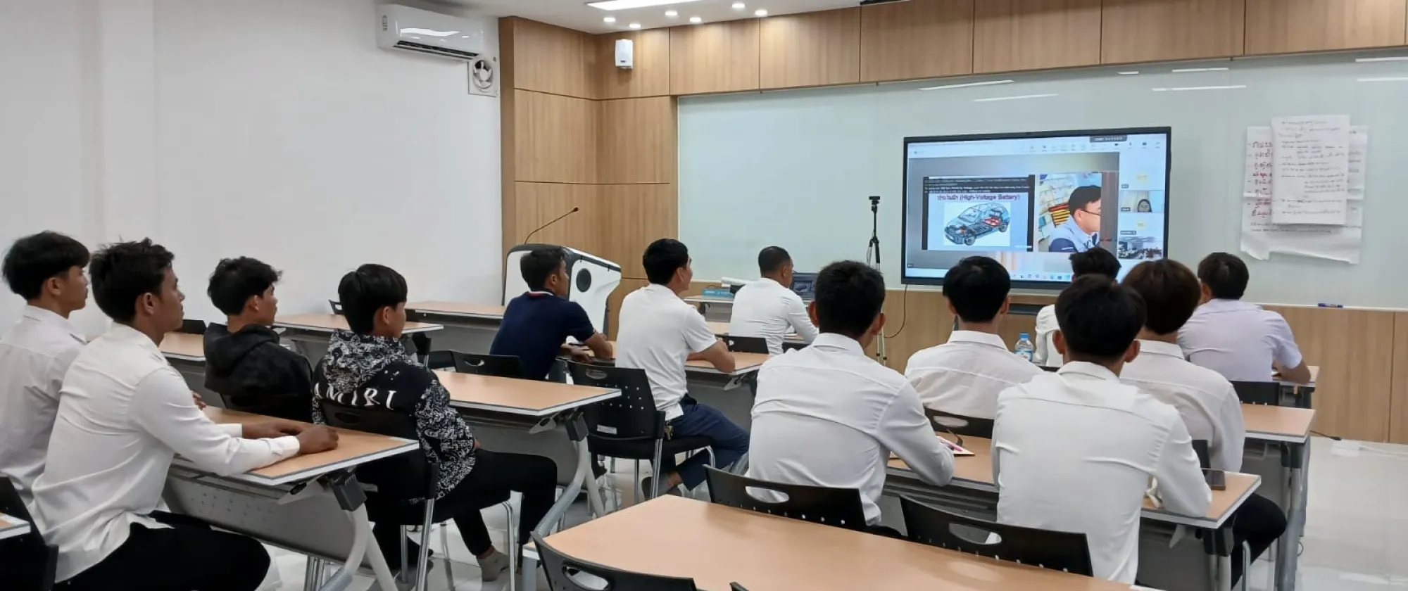 A group of students sits in a classroom watching an online training session displayed on a large screen.