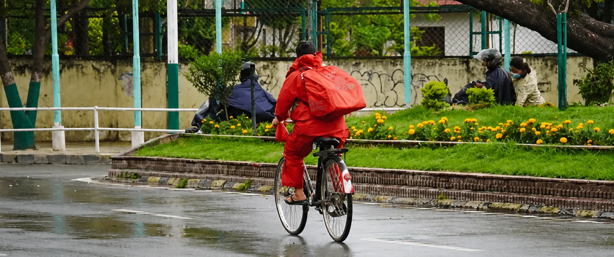 A bicycle delivery worker wearing a bright red rain suit and carrying a large insulated delivery bag rides along a rain-soaked street.