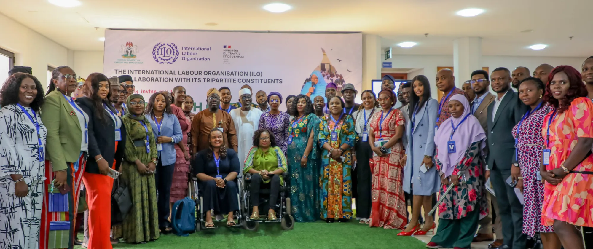 Dozens of participants stand together around senior officials for a group photograph in front of the event backdrop. The gathering includes representatives from government, workers' and employers' organizations, civil society and development partners.