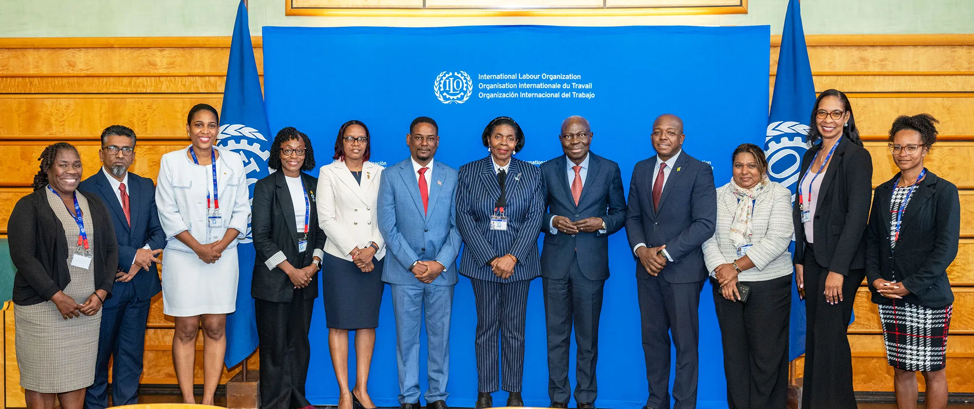 A formal group portrait of eleven delegates standing in a row in front of a bright blue International Labour Organization (ILO) backdrop. They are flanked by two ILO flags and posed against a wooden wall paneled room.