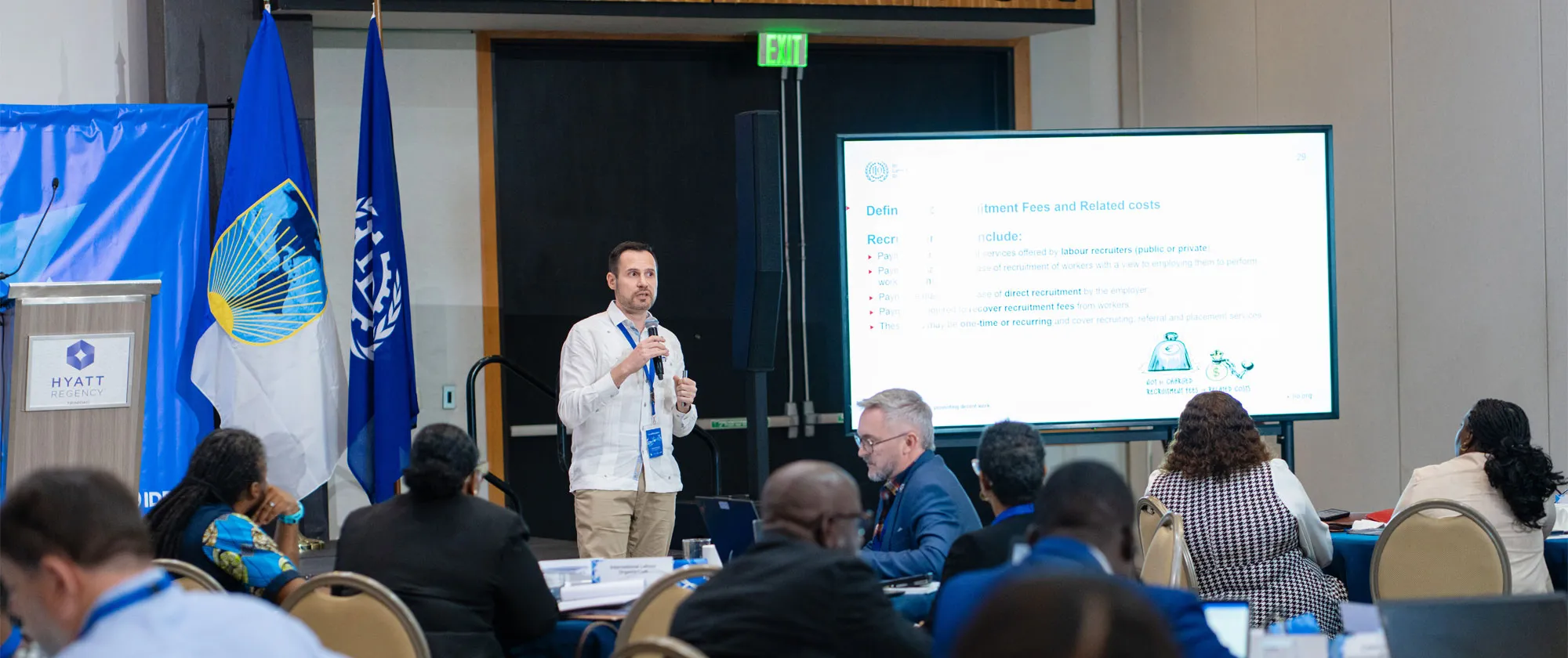 A male presenter speaking into a microphone during a conference presentation at a Hyatt Regency venue.