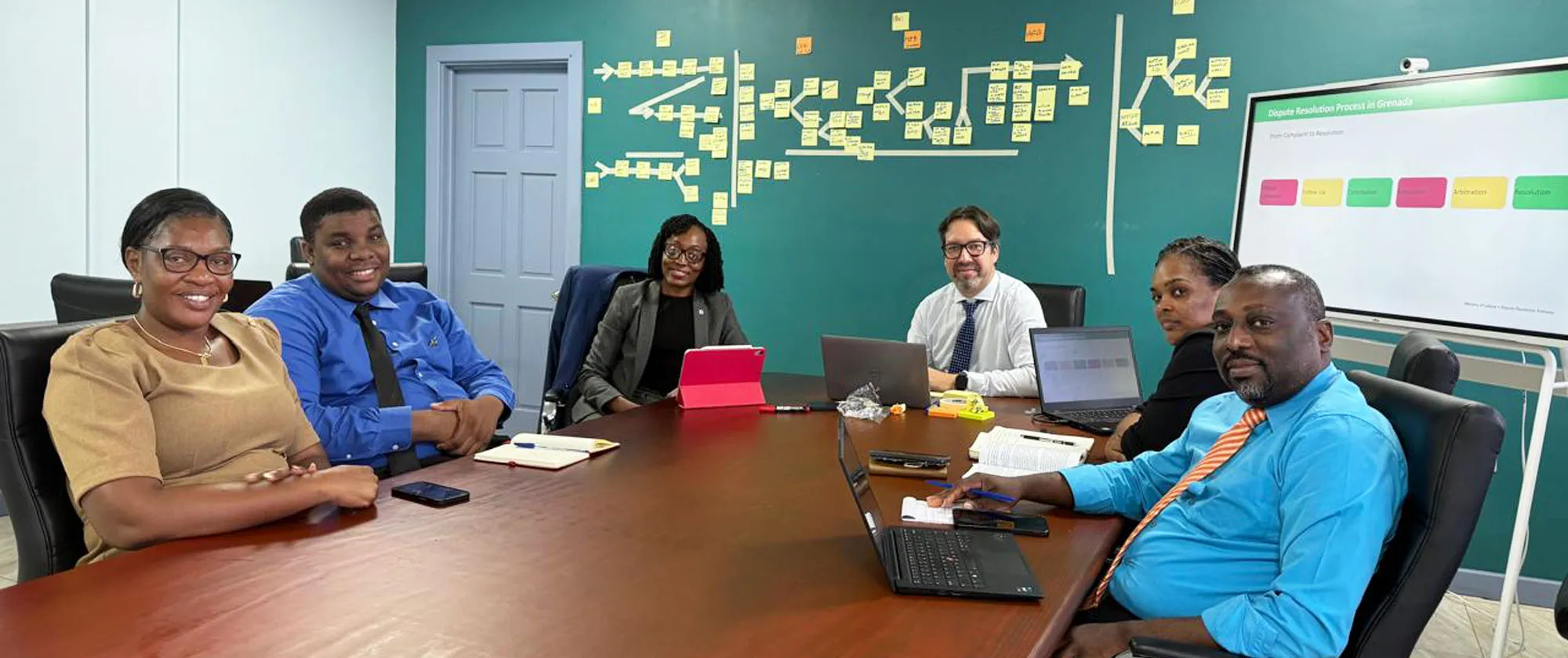 A group of six professionals sits around a large wooden conference table in a meeting room.
