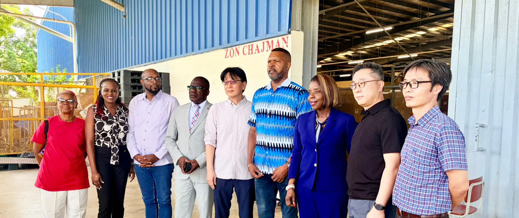 “A group of ten adults stand side by side inside an industrial facility with a high metal roof and blue corrugated walls. They are dressed in a mix of business and smart-casual attire and appear to be part of an official visit or mission.