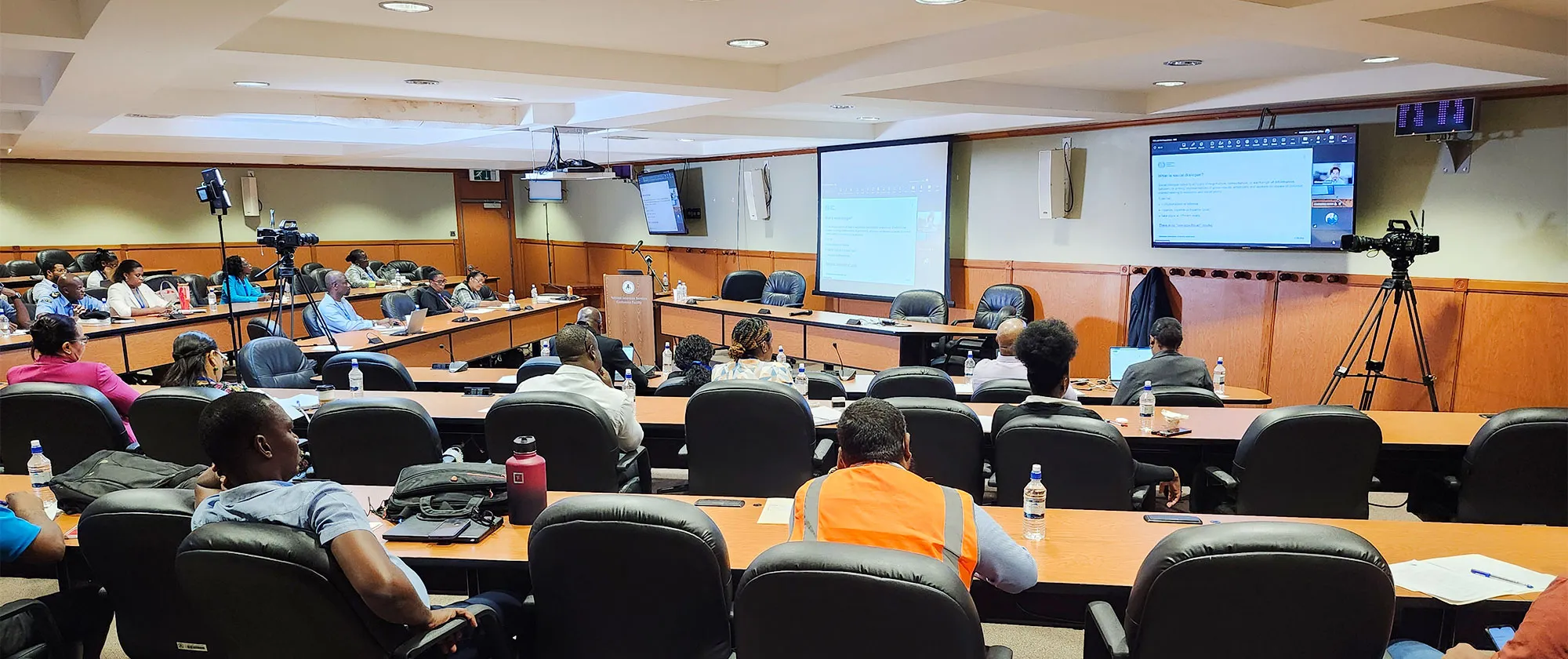A wide, high-angle shot of a hybrid workshop or parliamentary-style committee room. Participants are seated at long, curved tiered wooden desks facing a presentation area equipped with projection screens and a large digital monitor