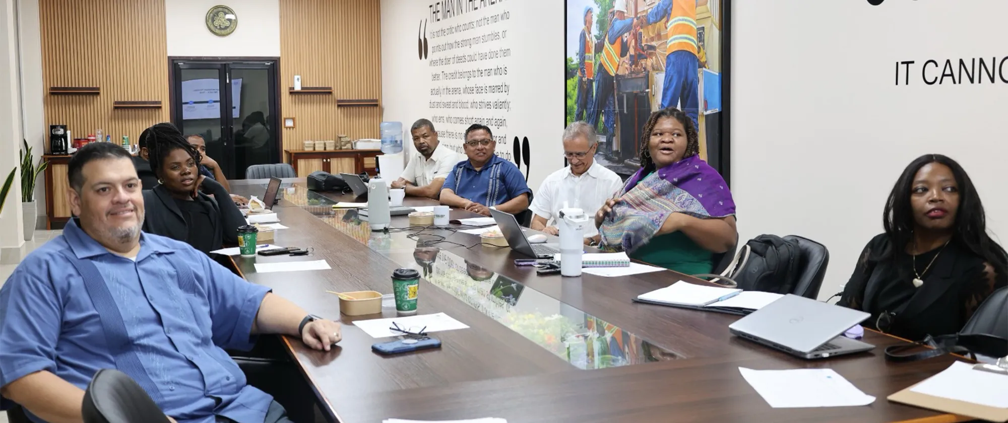 A group of professionals sits along a long, modern conference table during a project steering committee meeting. A woman in a green top and purple shawl speaks actively while others listen.