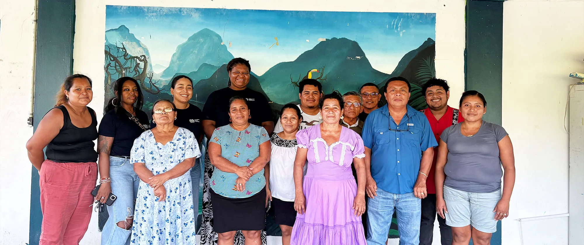 A group photo of thirteen individuals standing side-by-side outdoors against a painted mural backdrop depicting lush green mountains and a blue sky.