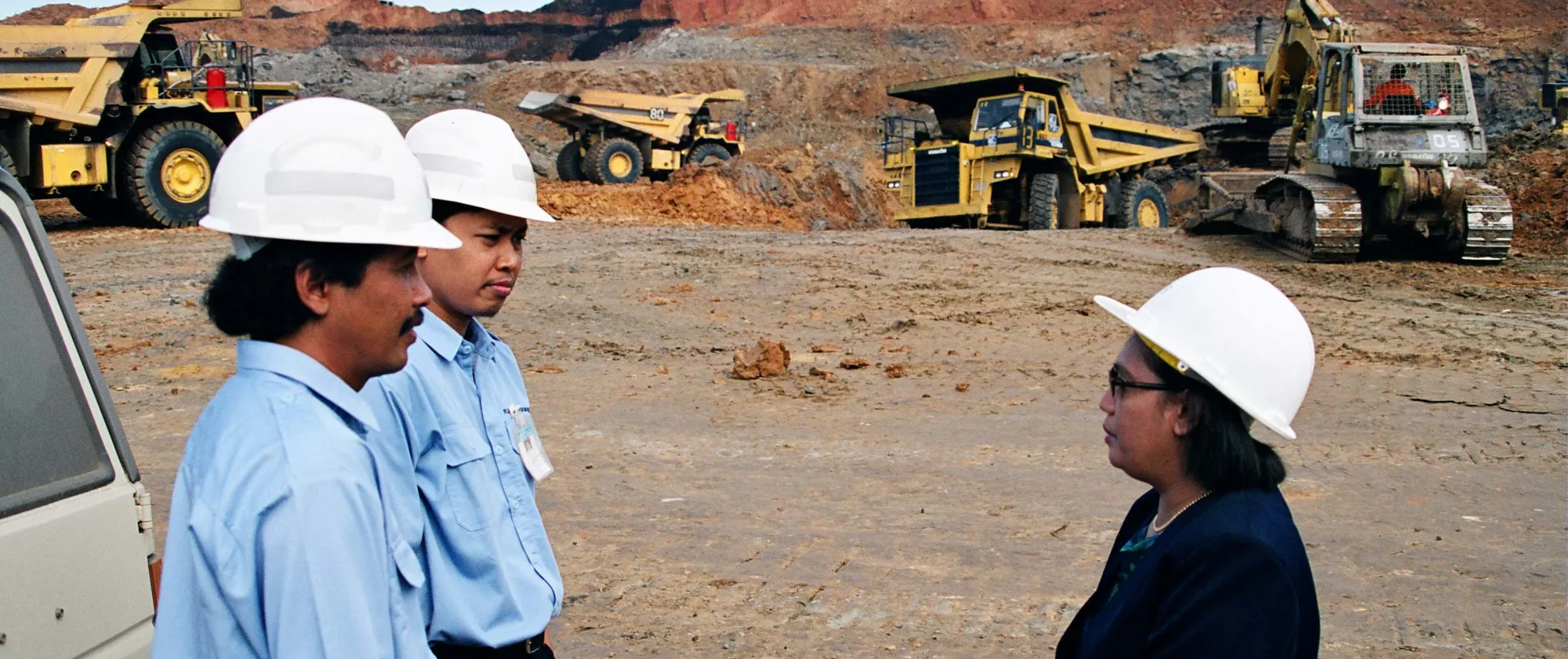 Three people wearing white hard hats stand in conversation at an open-pit mining site.