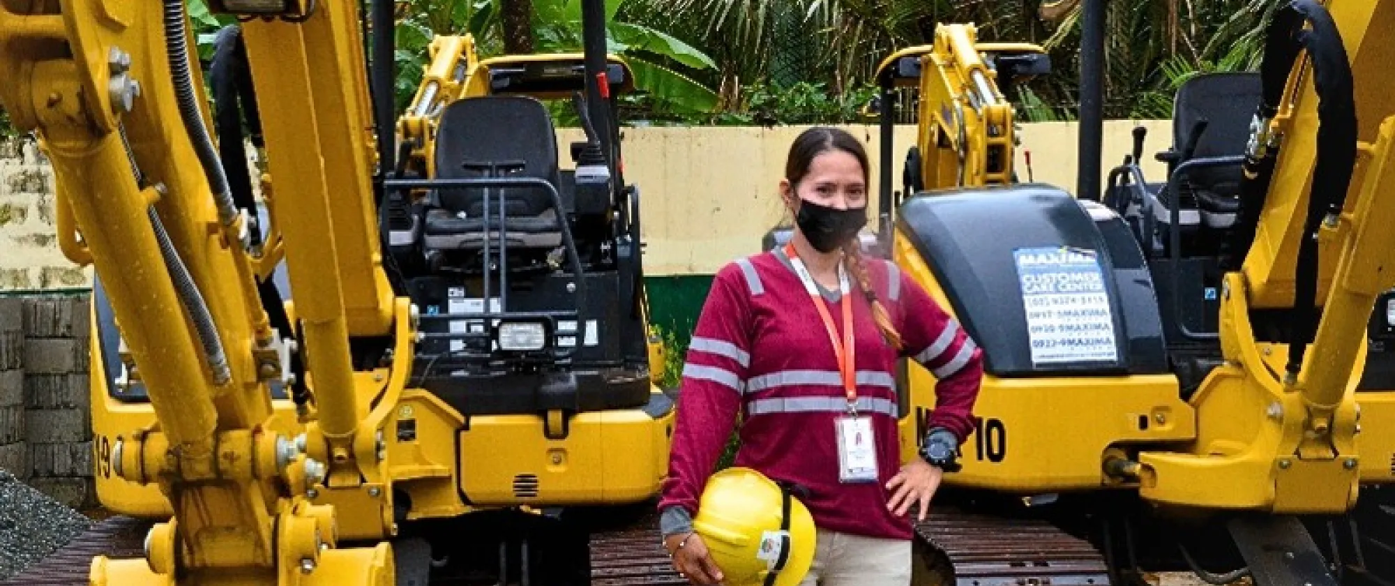 A woman wearing a high-visibility work shirt, face mask, and identification badge stands between two mini excavators while holding a yellow safety helmet.