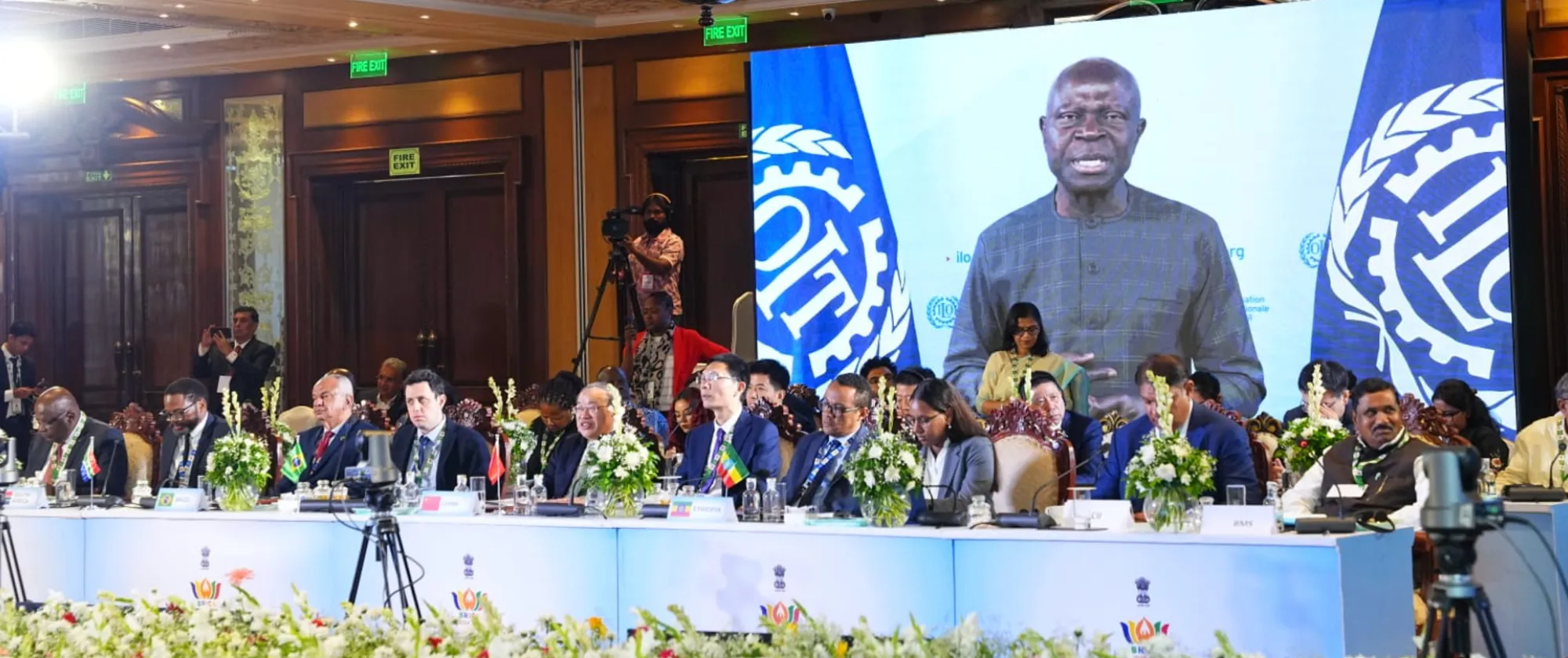 Participants sit at a long table during a conference with a large screen behind them displaying a speaker.