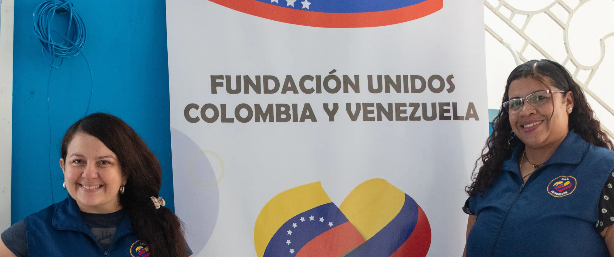Two female FUNCOLVEN staff members stand smiling beside a banner for Fundación Unidos Colombia y Venezuela, which features the organization's name, logo, and contact information. The photo appears to have been taken indoors during an organizational event or community activity.