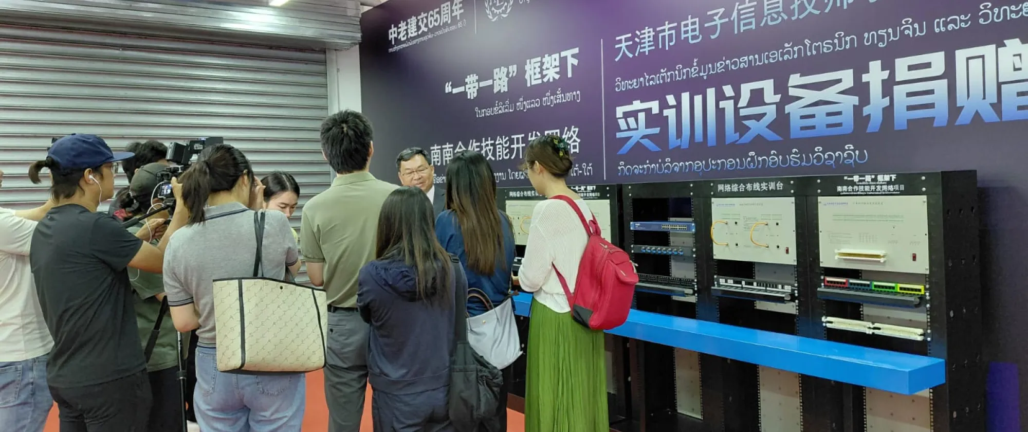 Several people including a video cameraman are speaking to some one. A large sign with Chinese text hangs on the wall.