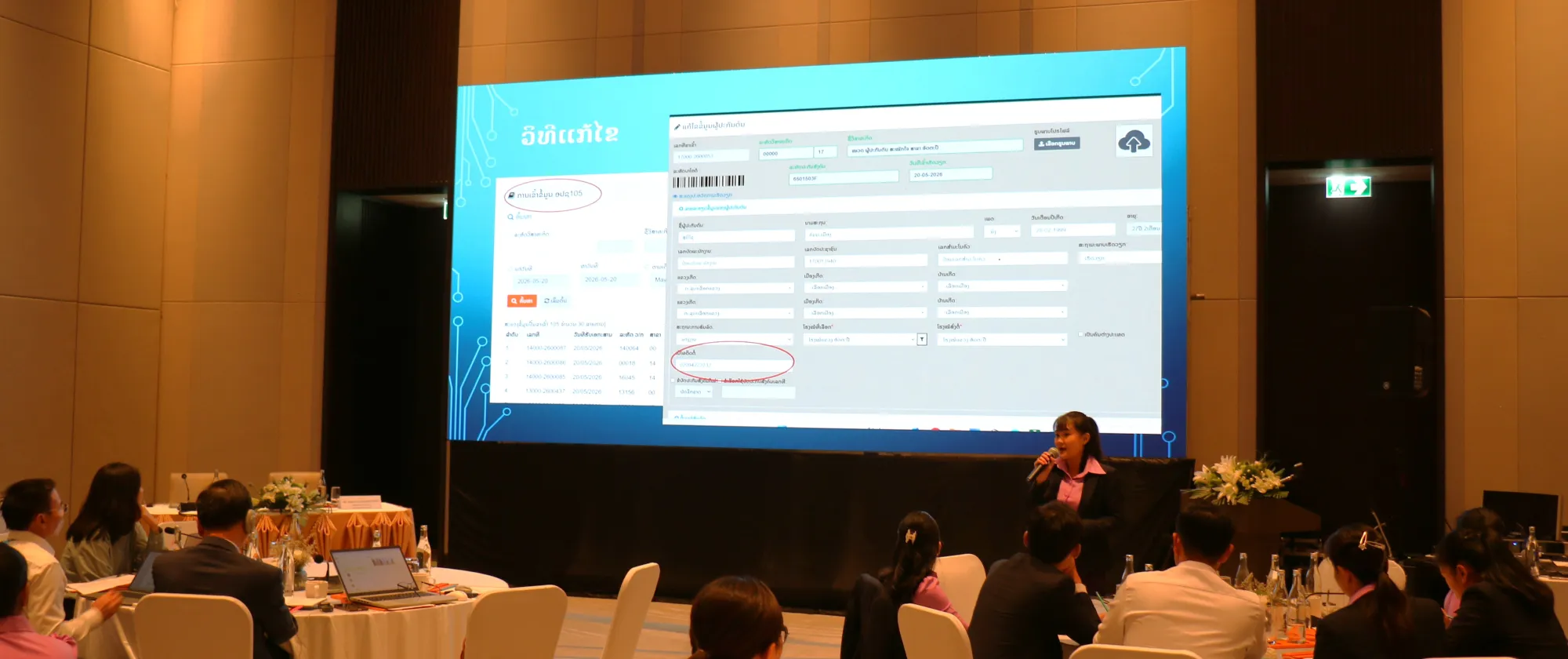 Participants attend a training session in a hotel meeting room, facing a large presentation screen at the front of the hall.