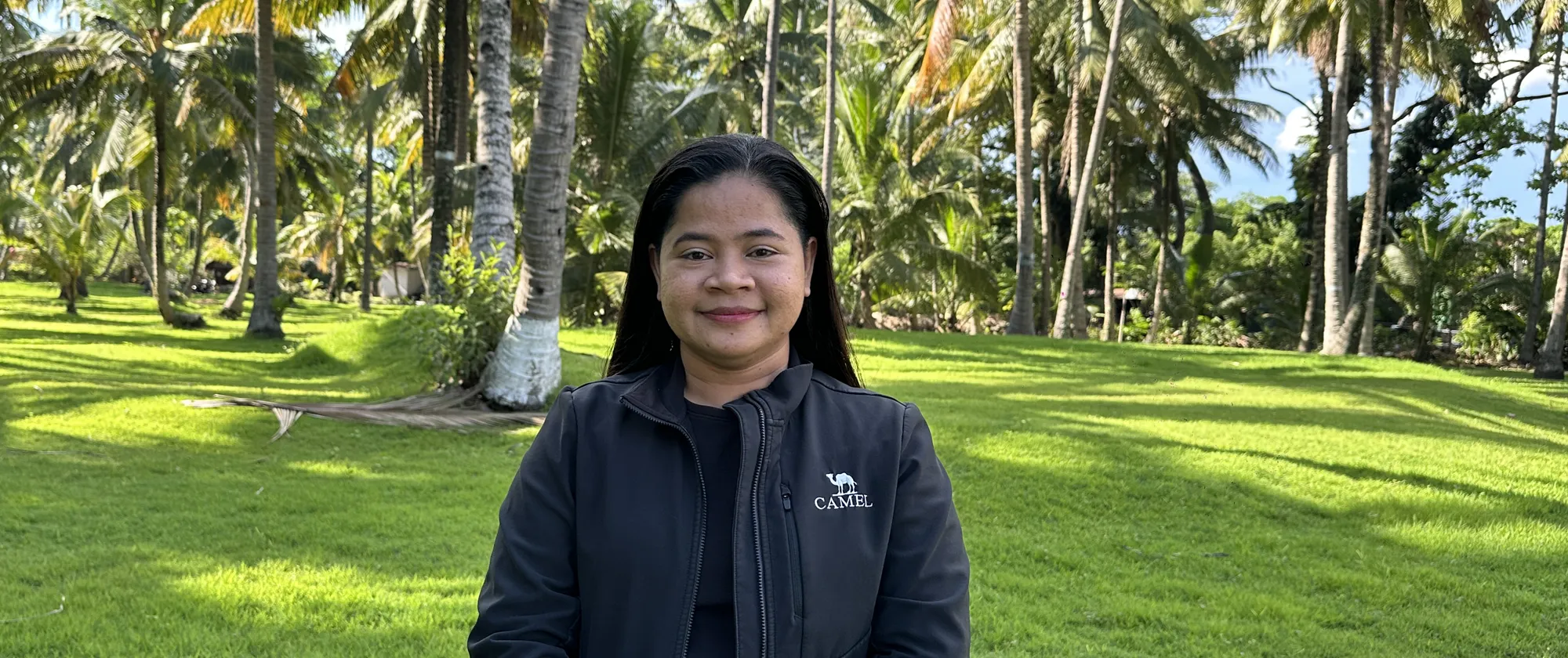 Melba wearing a black jacket and standing behind a green field with coconut trees