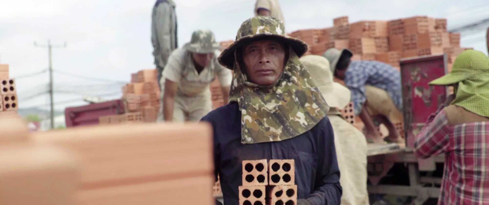 A construction worker wearing sun-protective clothing stands holding clay bricks.