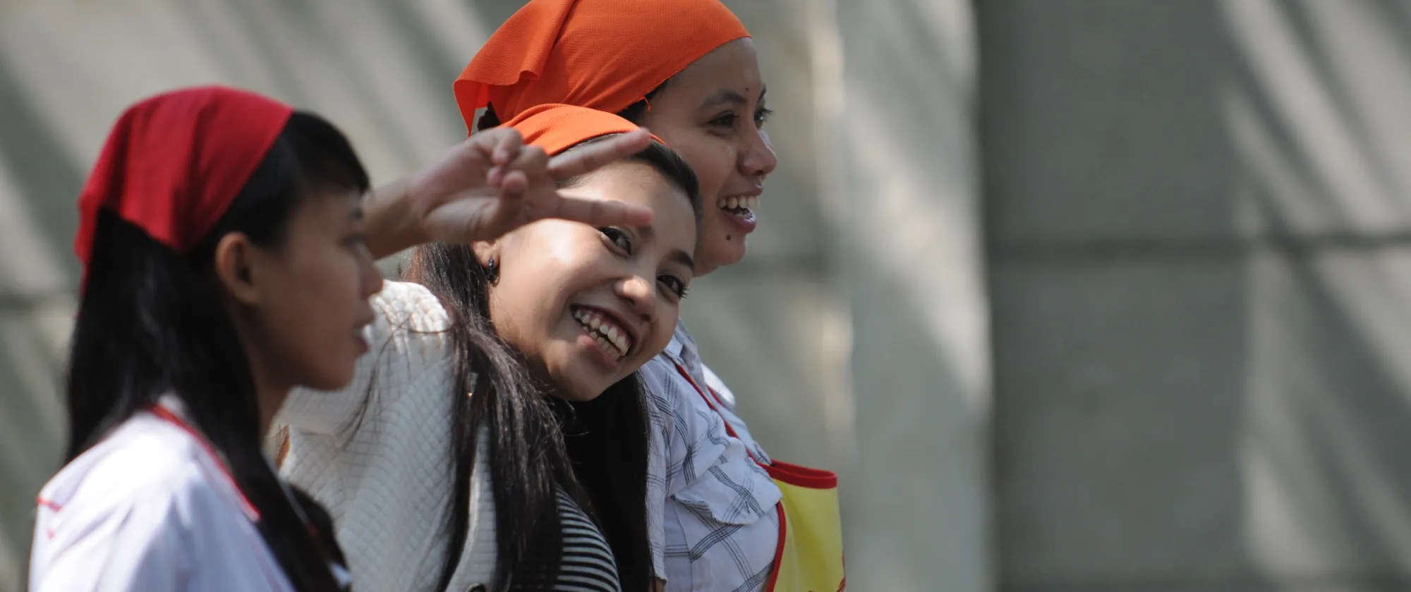 Tre giovani donne con foulard arancioni in piedi insieme all'aperto, sorridenti. Una fa il segno della pace, mentre le altre guardano davanti a sé.