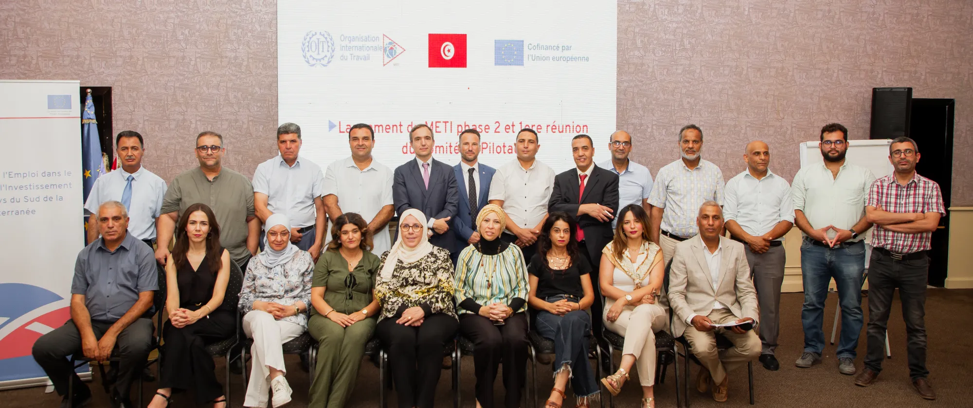 Photo de groupe des participants au lancement de la deuxième phase du programme METI en Tunisie et à la première réunion du comité de pilotage. Les participants posent devant un écran affichant le titre de l’événement ainsi que les logos des partenaires.
