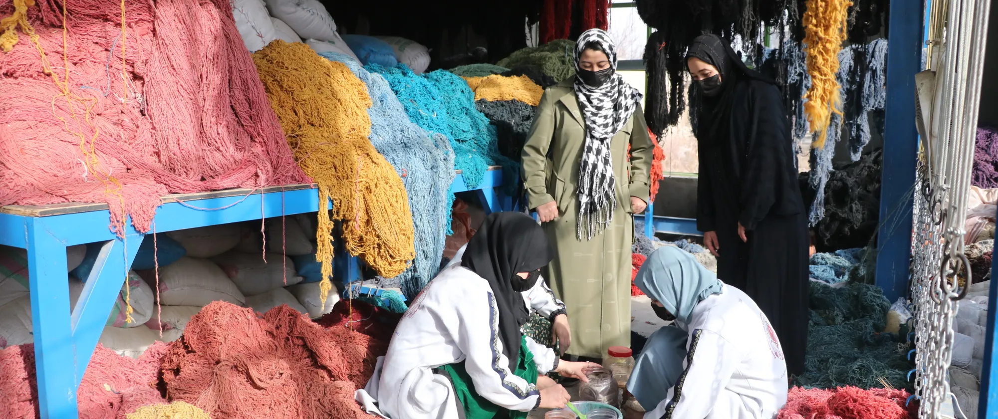 Mujeres trabajan entre grandes madejas de hilos de colores, mientras otras dos mujeres observan su trabajo.