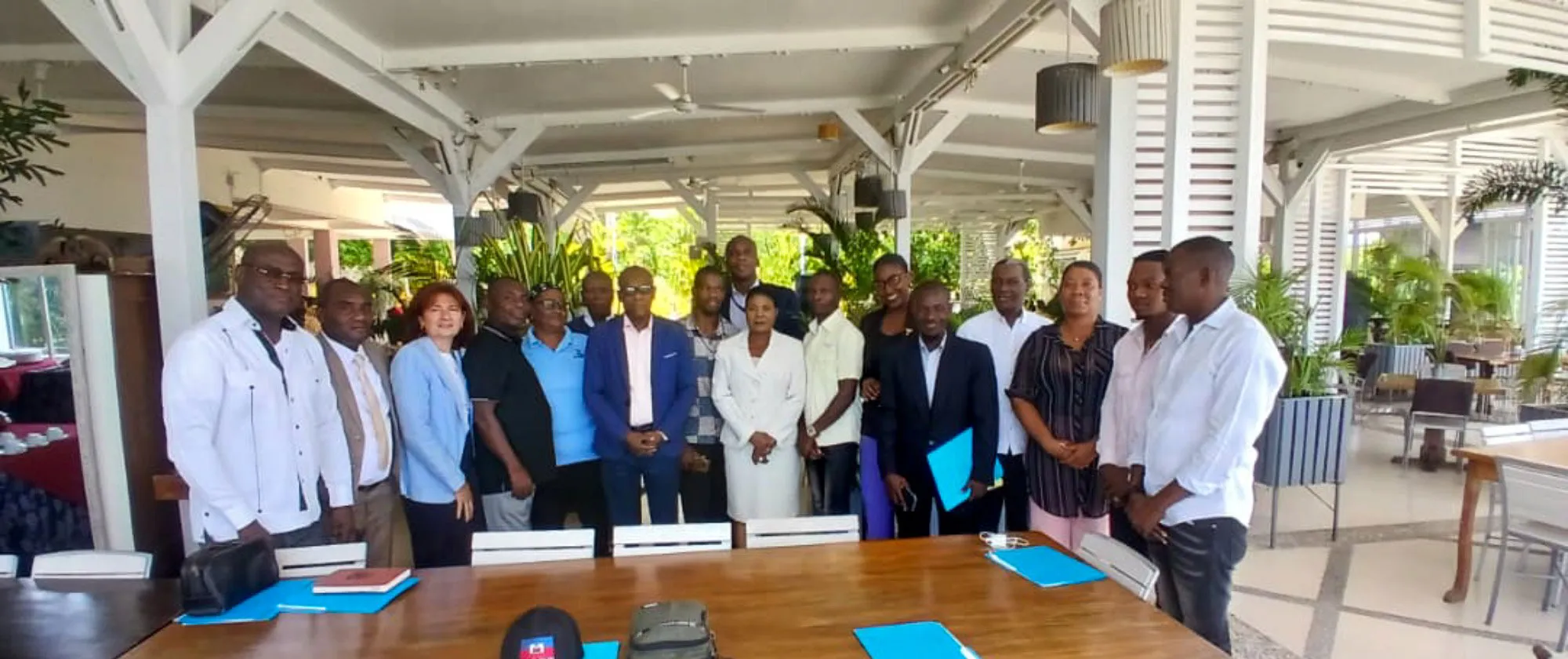 A group of about 15 men and women stand together behind a long wooden table during a meeting in a covered open-air venue with white wooden beams and lattice screens. Blue document folders and a cap featuring the Haitian flag sit on the table in the foreground.