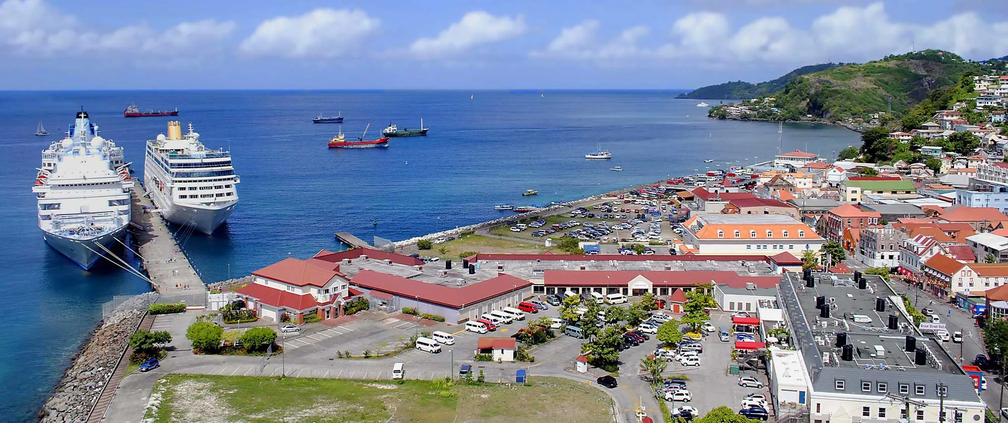 High-angle wide view of St. George’s harbour in Grenada, featuring two large cruise ships docked along the pier in clear blue water. Red-roofed port buildings, parking areas, and the colorful town stretch along the bay toward lush green hills under a bright sky.