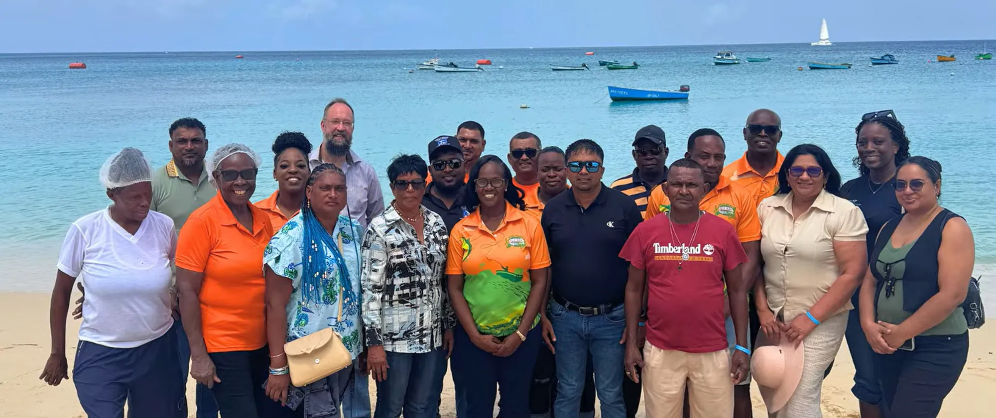 A group of fisheries knowledge-exchange participants standing together on a sandy beach, with bright blue ocean waters and small fishing boats behind them.