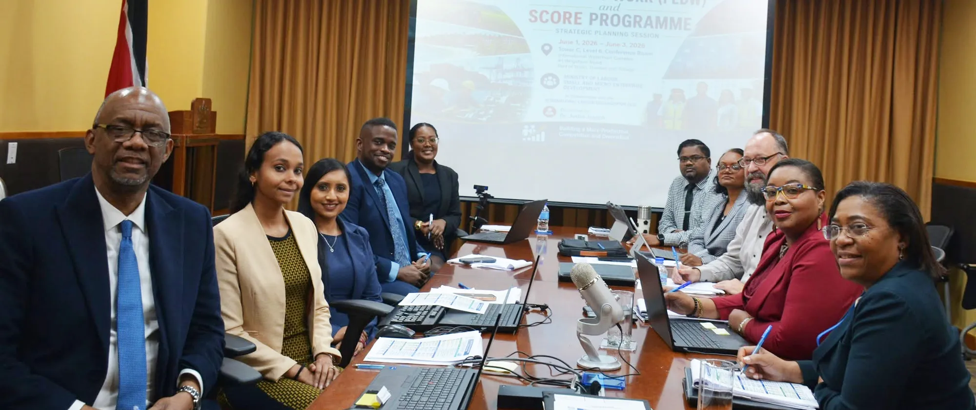 Participants seated around a conference table during a Strategic Planning Session, with a presentation screen and the Trinidad and Tobago national flag in the background.