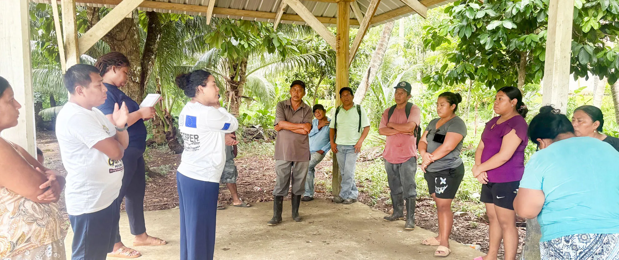A group of community members gathers under an open-air wooden shelter for a discussion led by a facilitator wearing an EU-ILO PROSPER long-sleeve shirt.
