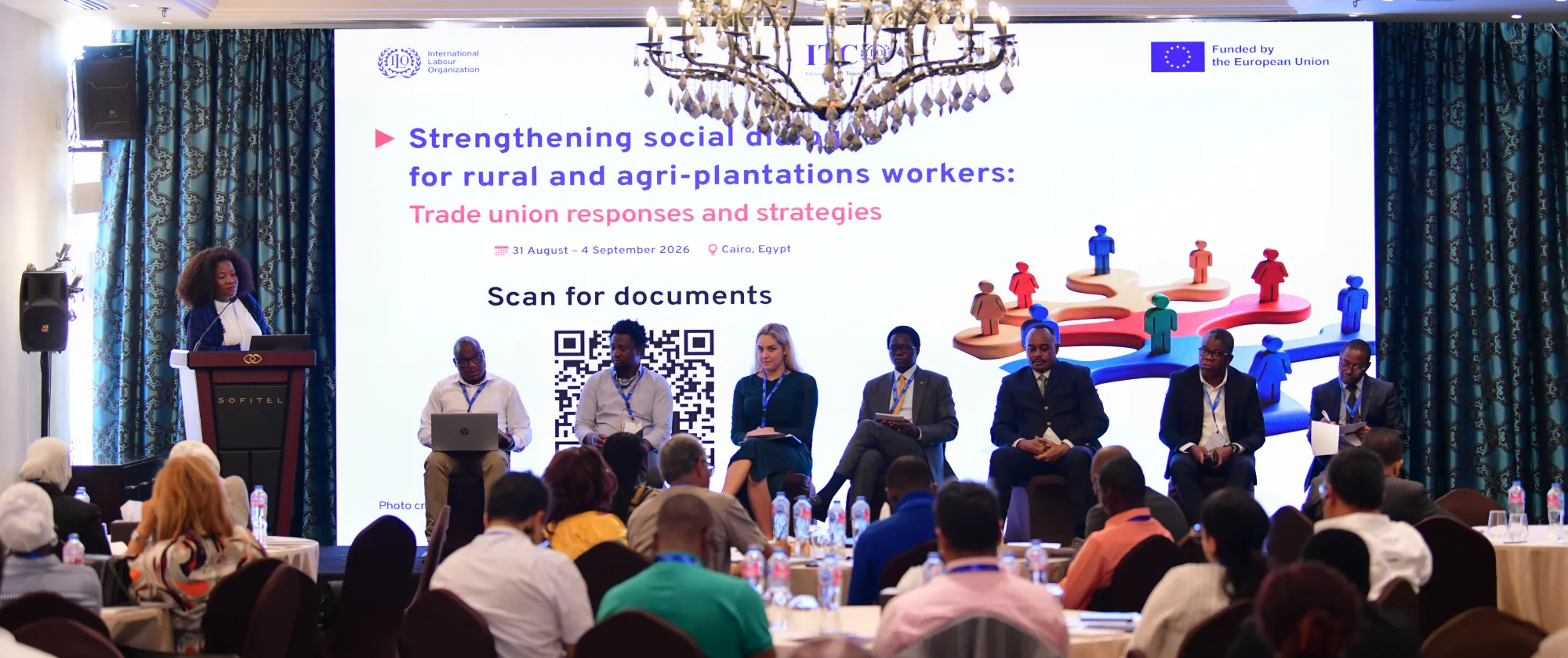 A speaker addresses participants seated in a conference room, with six panellists seated on stage in front of the event screen.