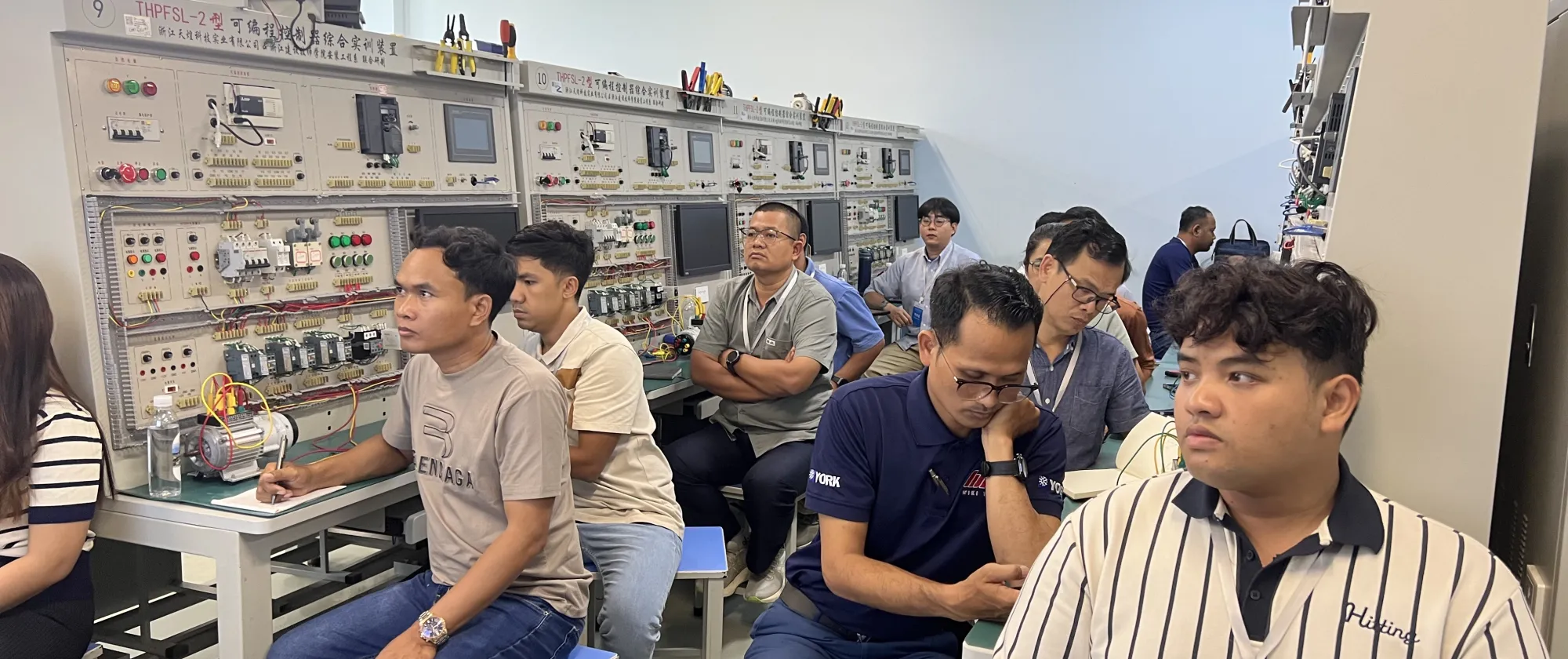 Several people sit at workbenches in a technical classroom, with shelves of electronic equipment behind them.