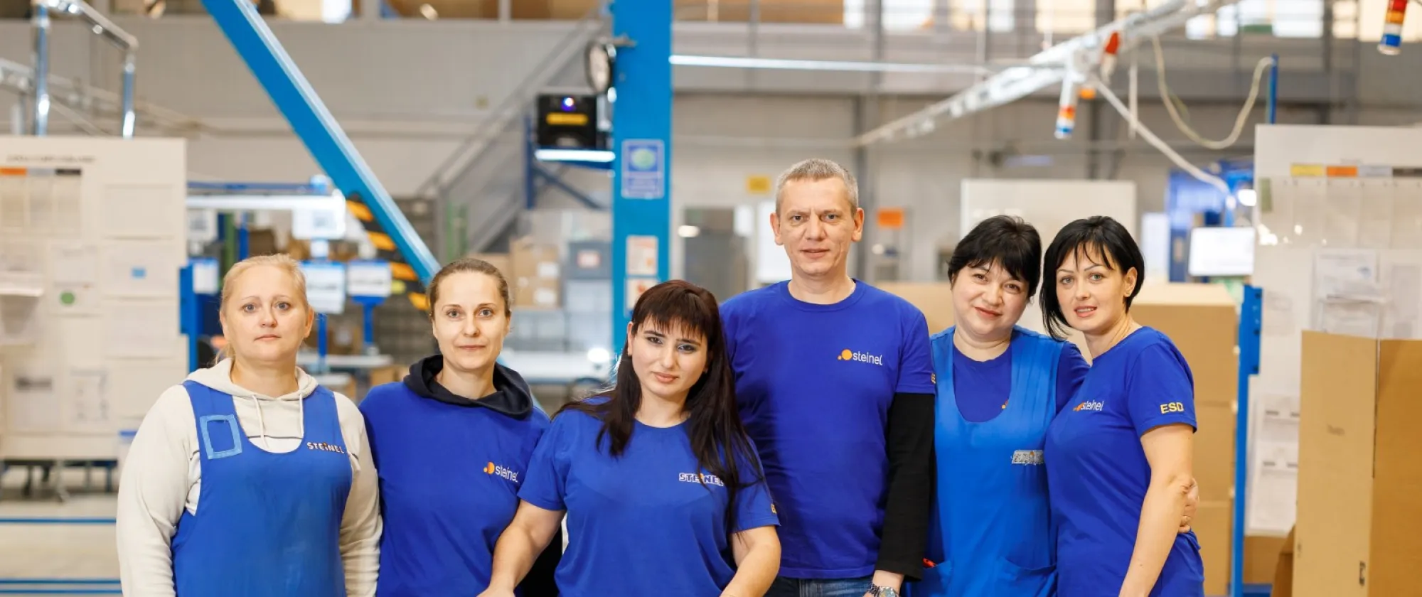 One man and six women workers, including a woman using a walker, stand together on a factory production floor.