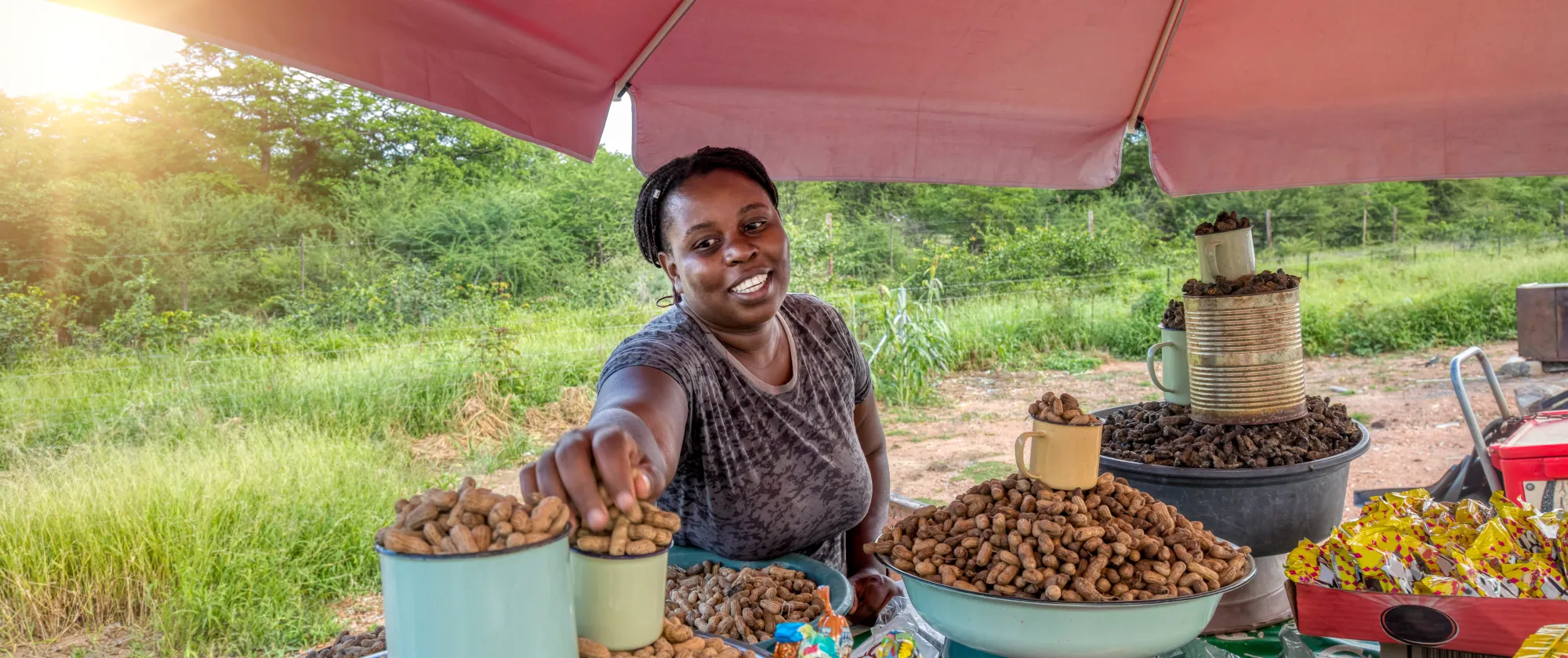 A smiling woman sells peanuts at an outdoor stall with lush green fields in the background