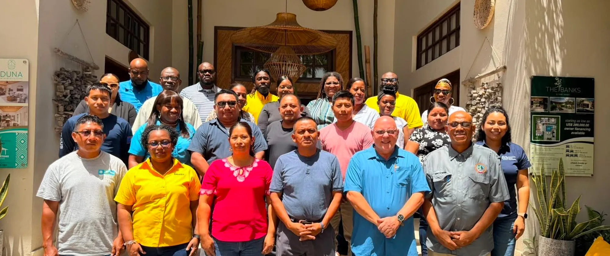 A large group photograph of approximately 25 to 30 people posing outside the entrance of a building. The diverse group of men and women are dressed in casual and business-casual attire, including brightly coloured t-shirts, polo shirts, and button-downs, and are organized in multiple rows facing the camera.