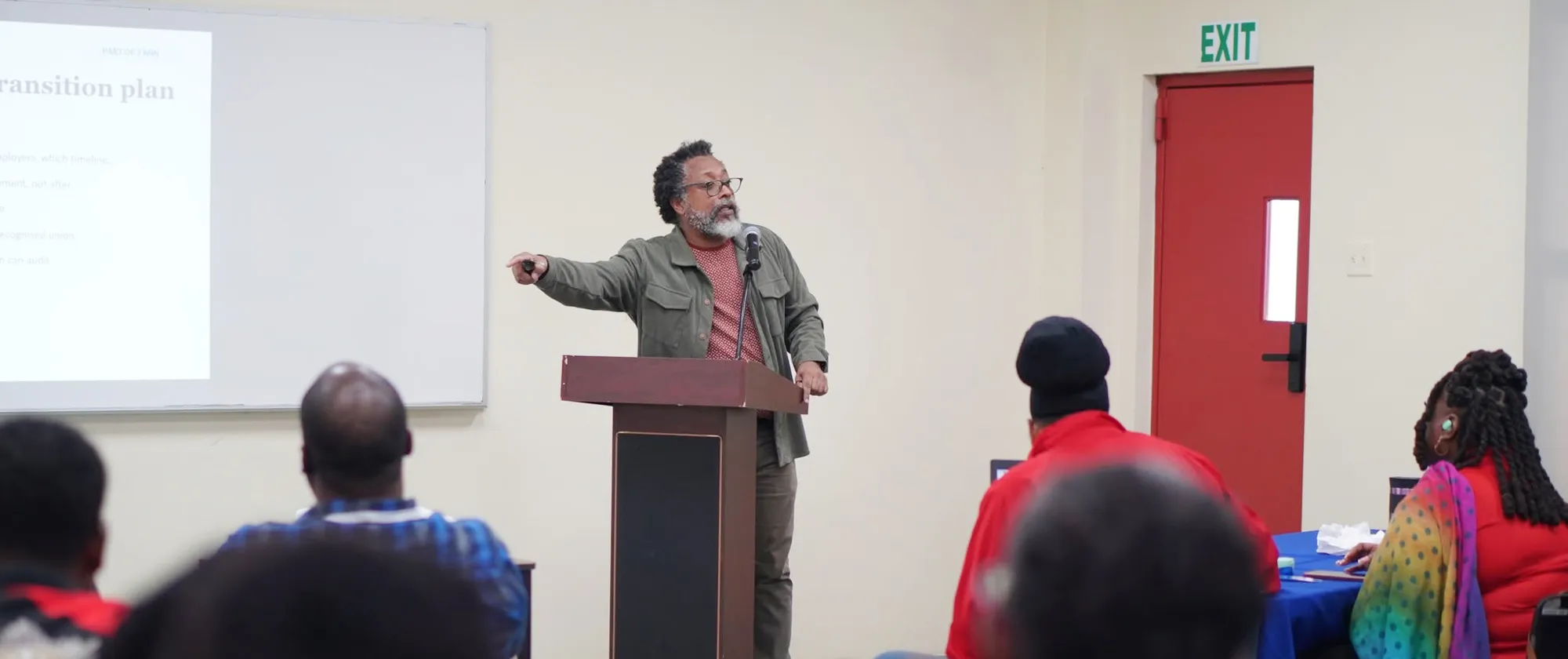 A photograph of a presenter addressing an audience in a bright training room. The speaker, a man with a grey beard and glasses, stands behind a dark wooden podium equipped with a microphone.