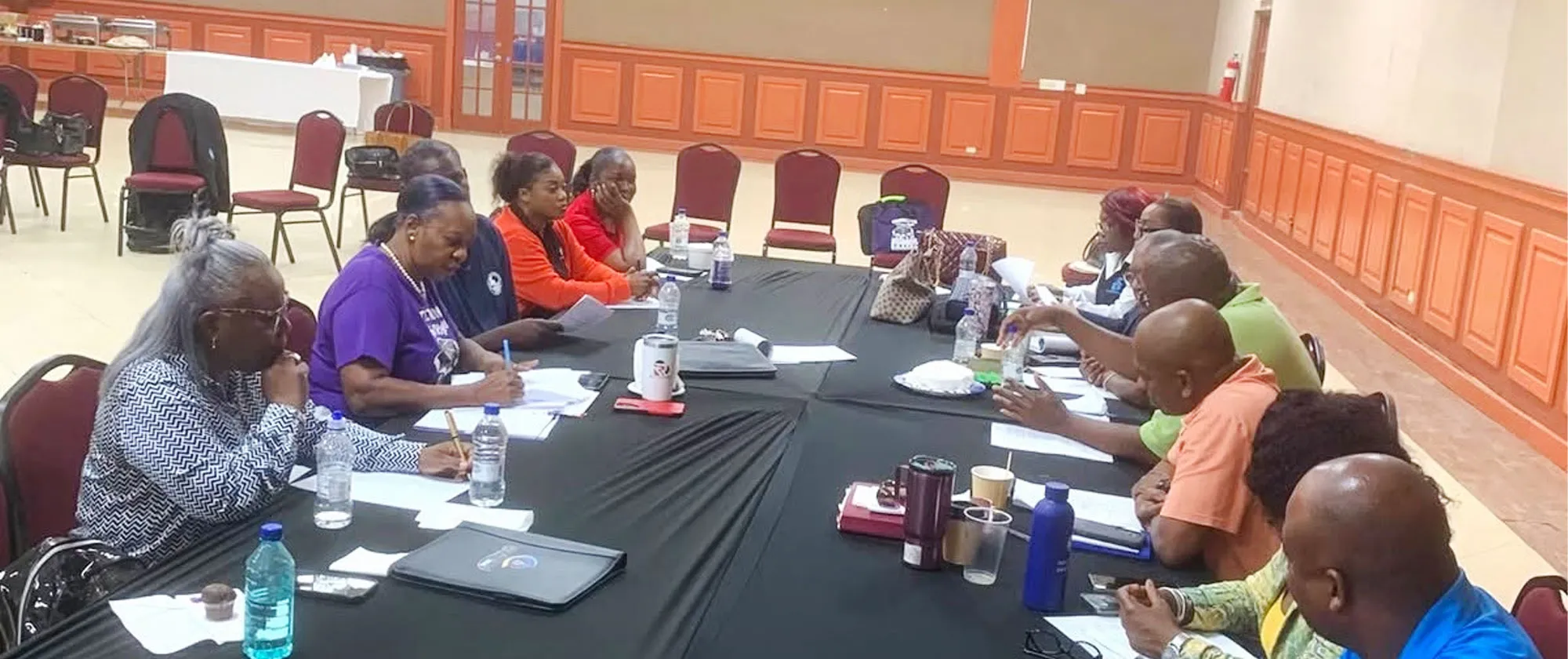 A photograph of approximately a dozen individuals participating in a meeting or training session. The attendees are seated on opposite sides of tables organized in a large configuration and draped with black tablecloths.