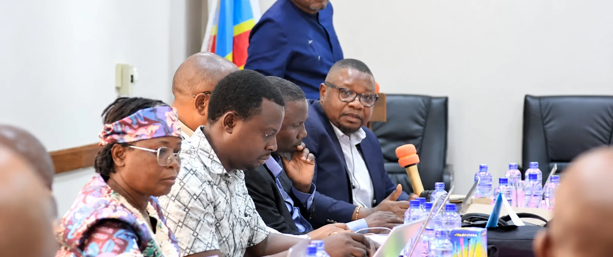 Des participants à l’atelier sur la protection sociale, assis autour d’une table de réunion à Kinshasa, écoutent les échanges et travaillent sur leurs ordinateurs portables.