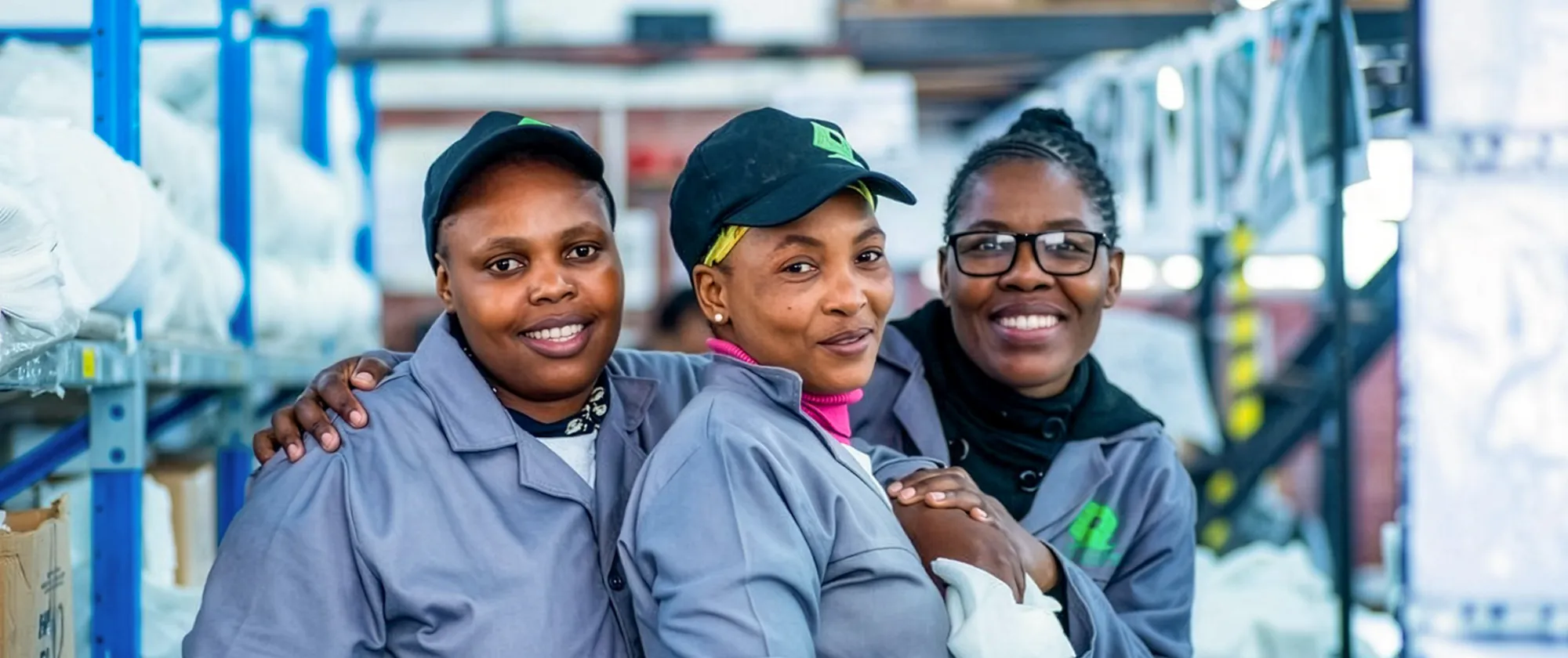 Three women factory workers in grey uniforms smile together during an ILO project field visit in South Africa in 2023.