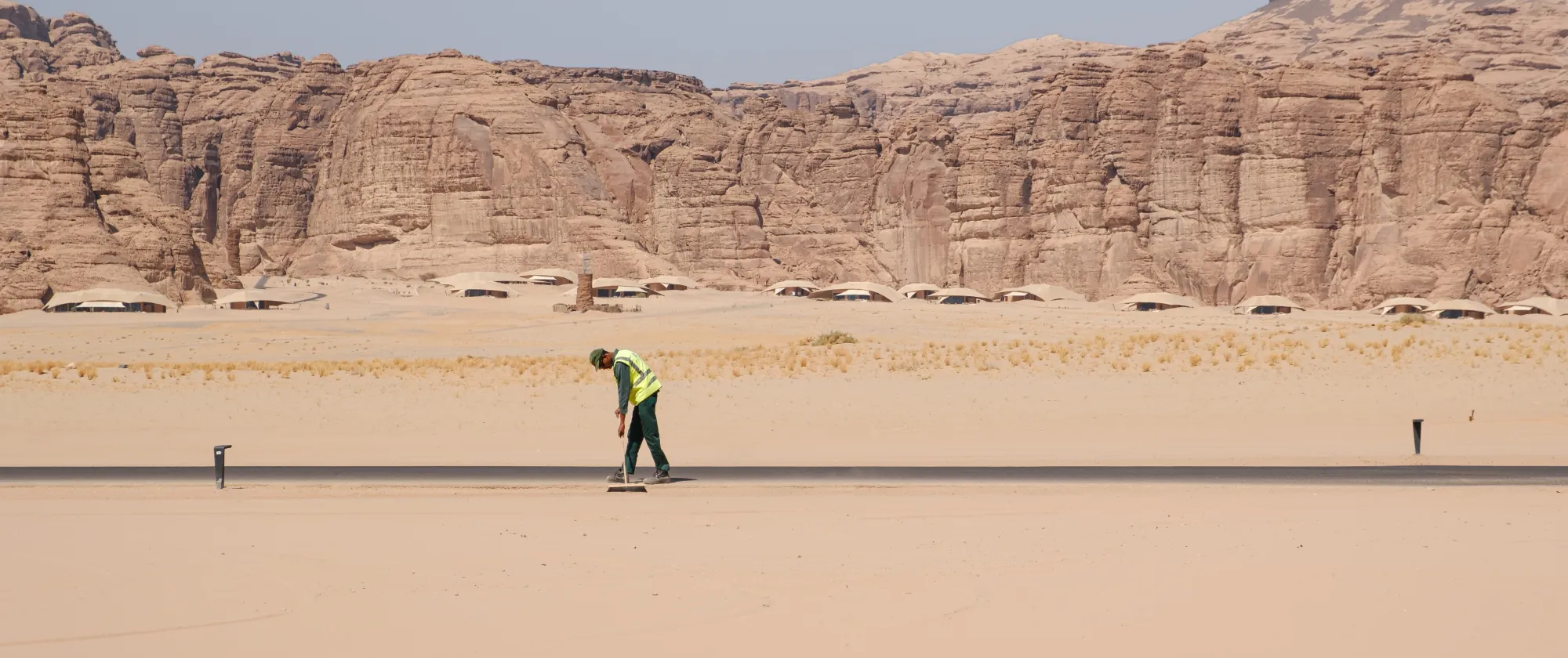 A worker wearing a high-visibility vest sweeps a paved road in a vast desert landscape, with towering sandstone cliffs in the background.