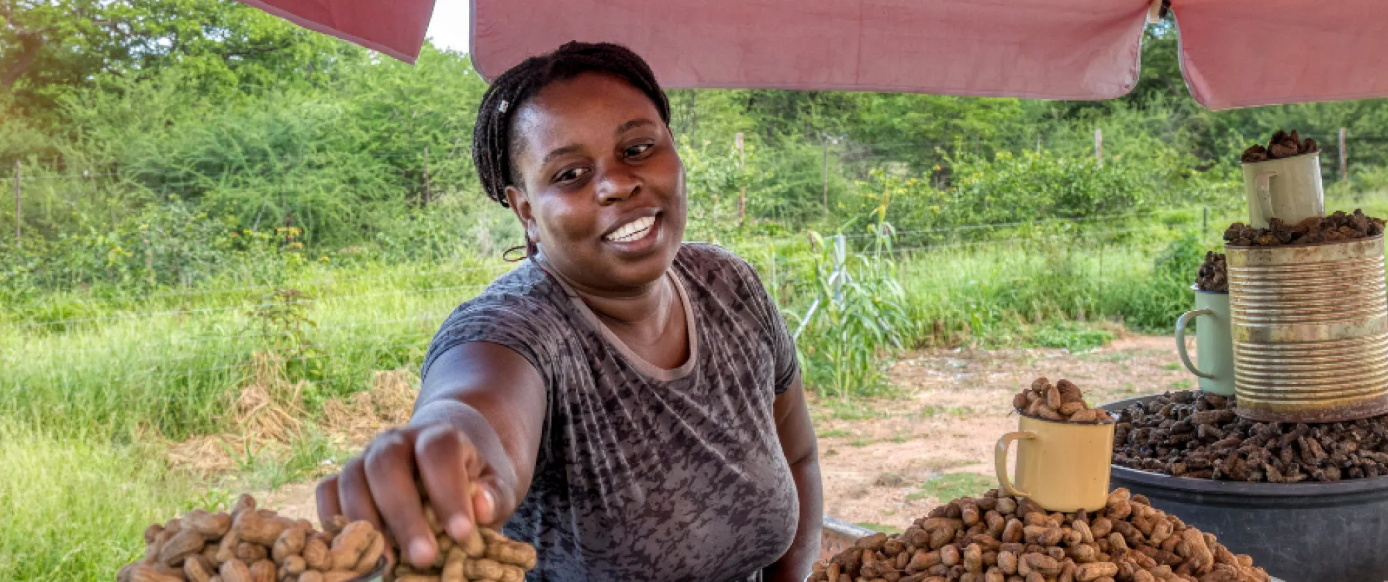 A smiling woman sells peanuts at an outdoor stall with lush green fields in the background