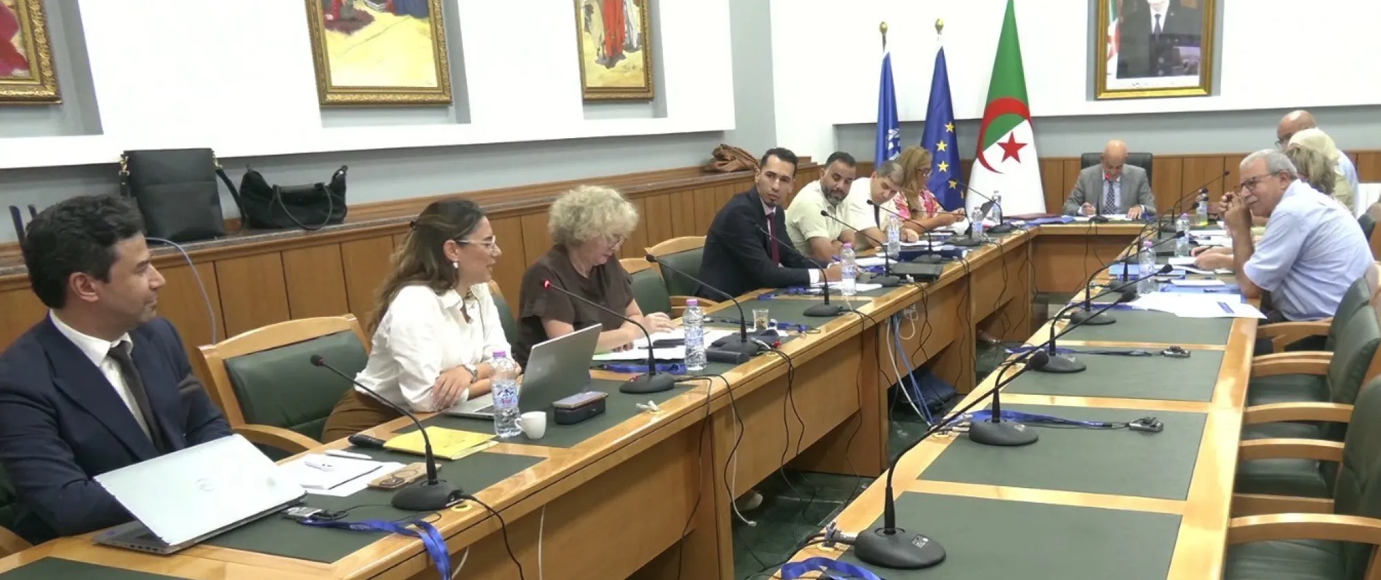 Vue d’ensemble d’une réunion réunissant plusieurs participants autour d’une grande table équipée de microphones et de documents. 