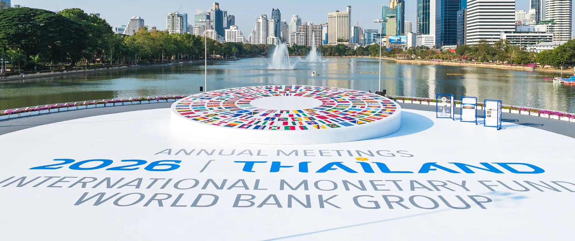 View of Bangkok’s skyline and a lake, with a circular display of international flags and signage for the 2026 IMF and World Bank Annual Meetings in the foreground.
