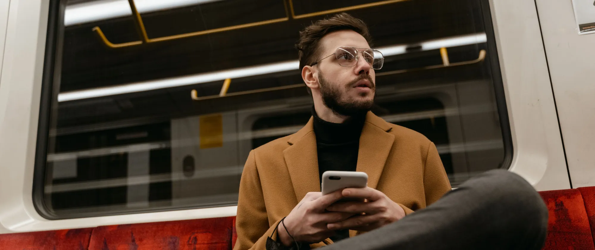 Casually dressed man with eyeglasses holds smartphone on a subway train seat.