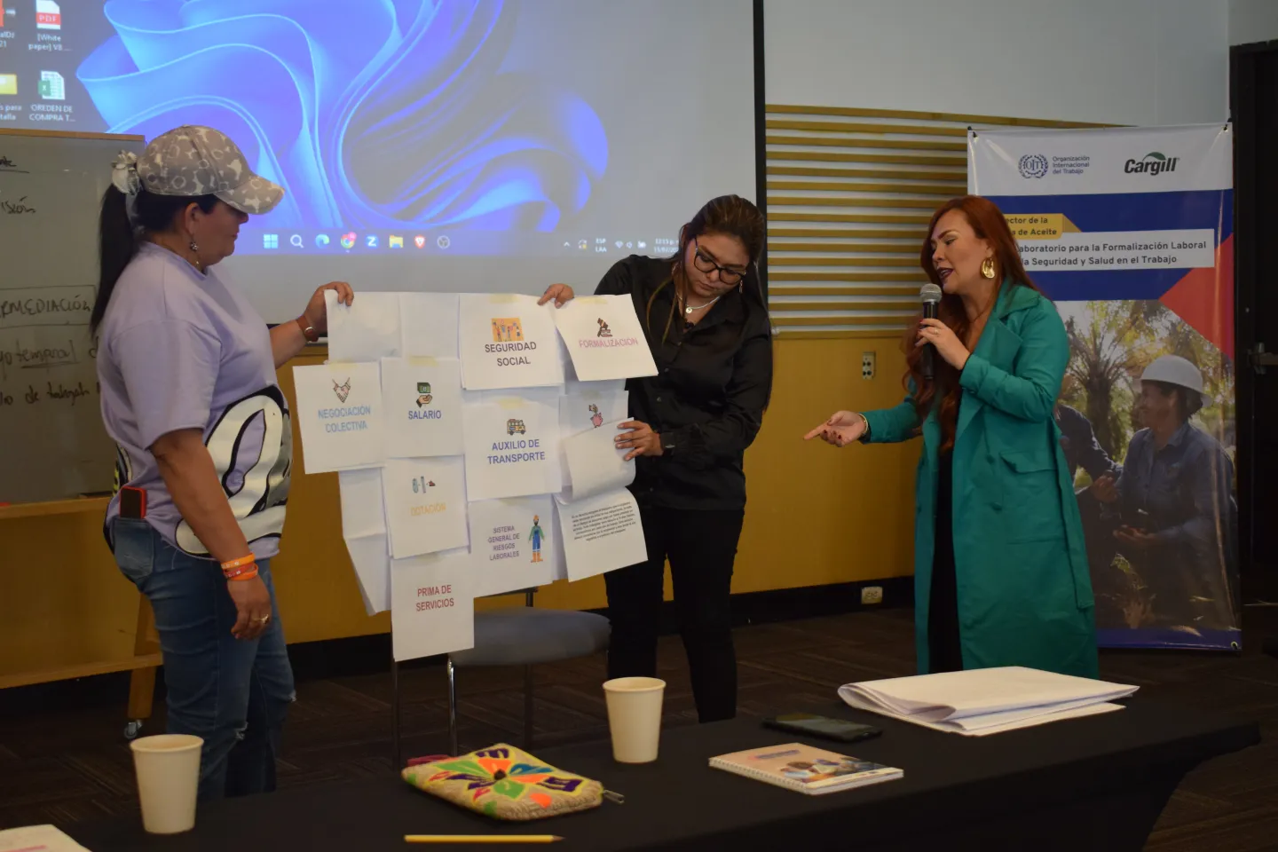 La expositora explicando los elementos clave del trabajo decente por medio de una cartelera sostenida por dos asistentes del evento