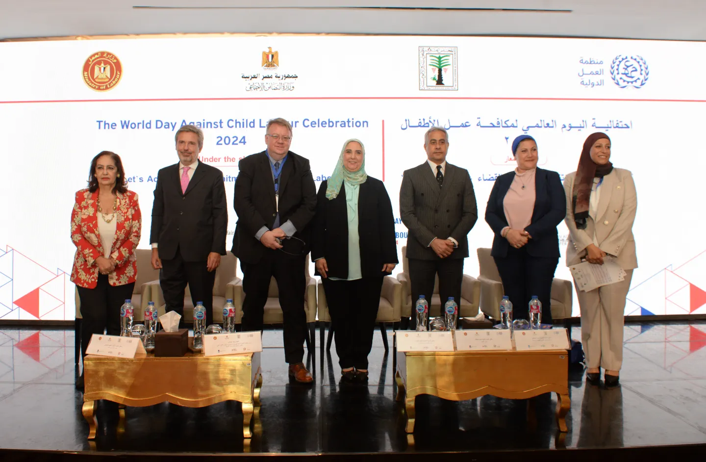 Representatives from the Government of Egypt and the ILO pose for a picture in the front of the room, some tables are in front of them and a big banner of the event behind.
