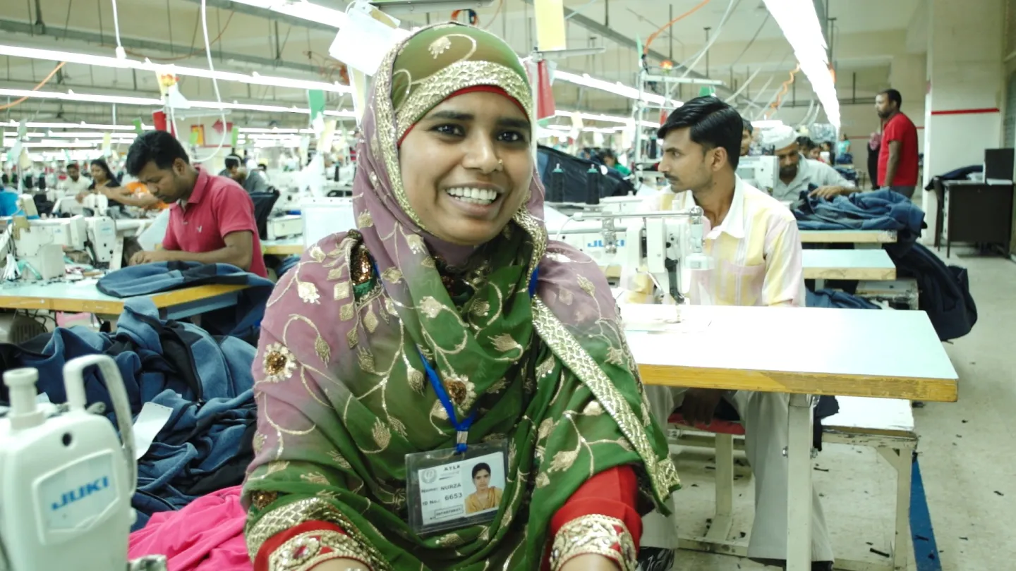 A Bangaldeshi woman at a garment workshop smiles with men in the background working, in Jordan