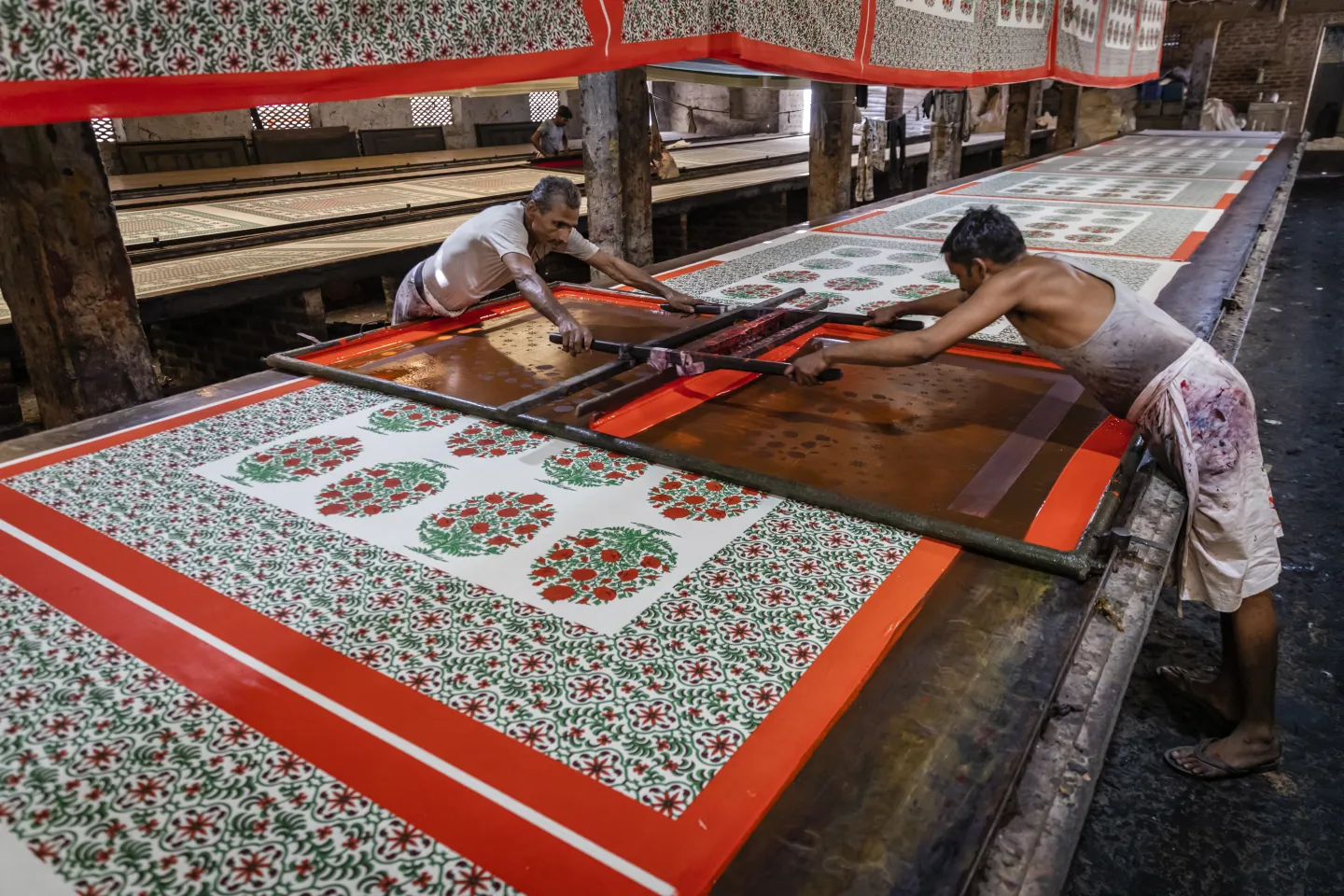 Two men working with a screen printing device on textile, the textile is red and green