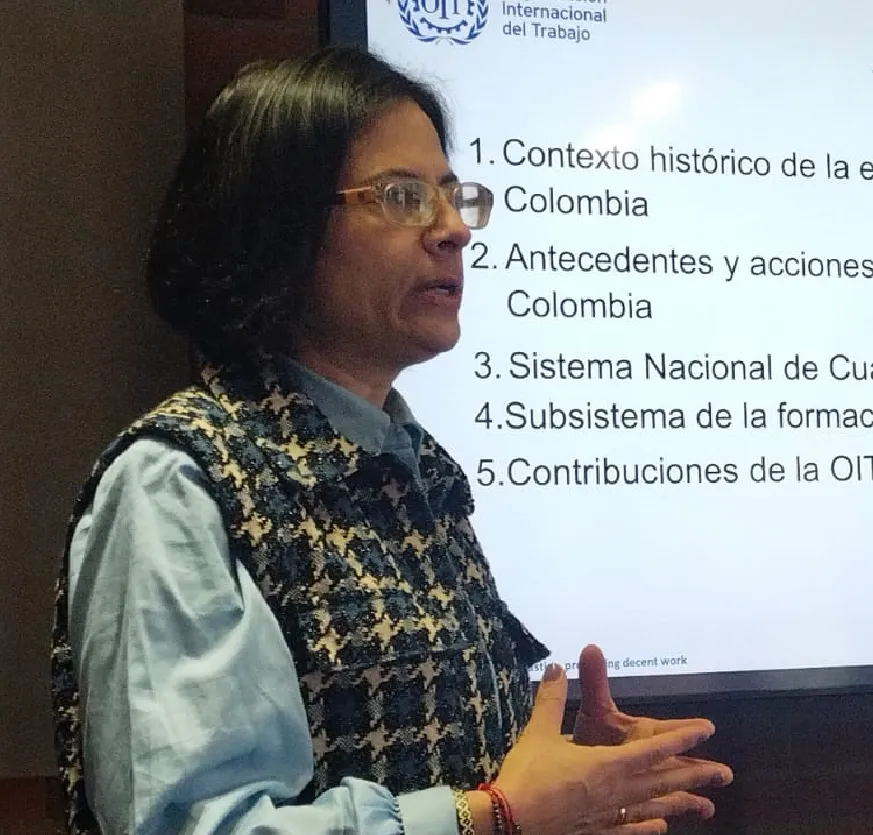Mujer hablando frente a la proyección de una presentación que muestra puntos sobre el contexto histórico y las contribuciones de la OIT en Colombia. Tiene el cabello corto, negro, lleva gafas y un chaleco estampado.