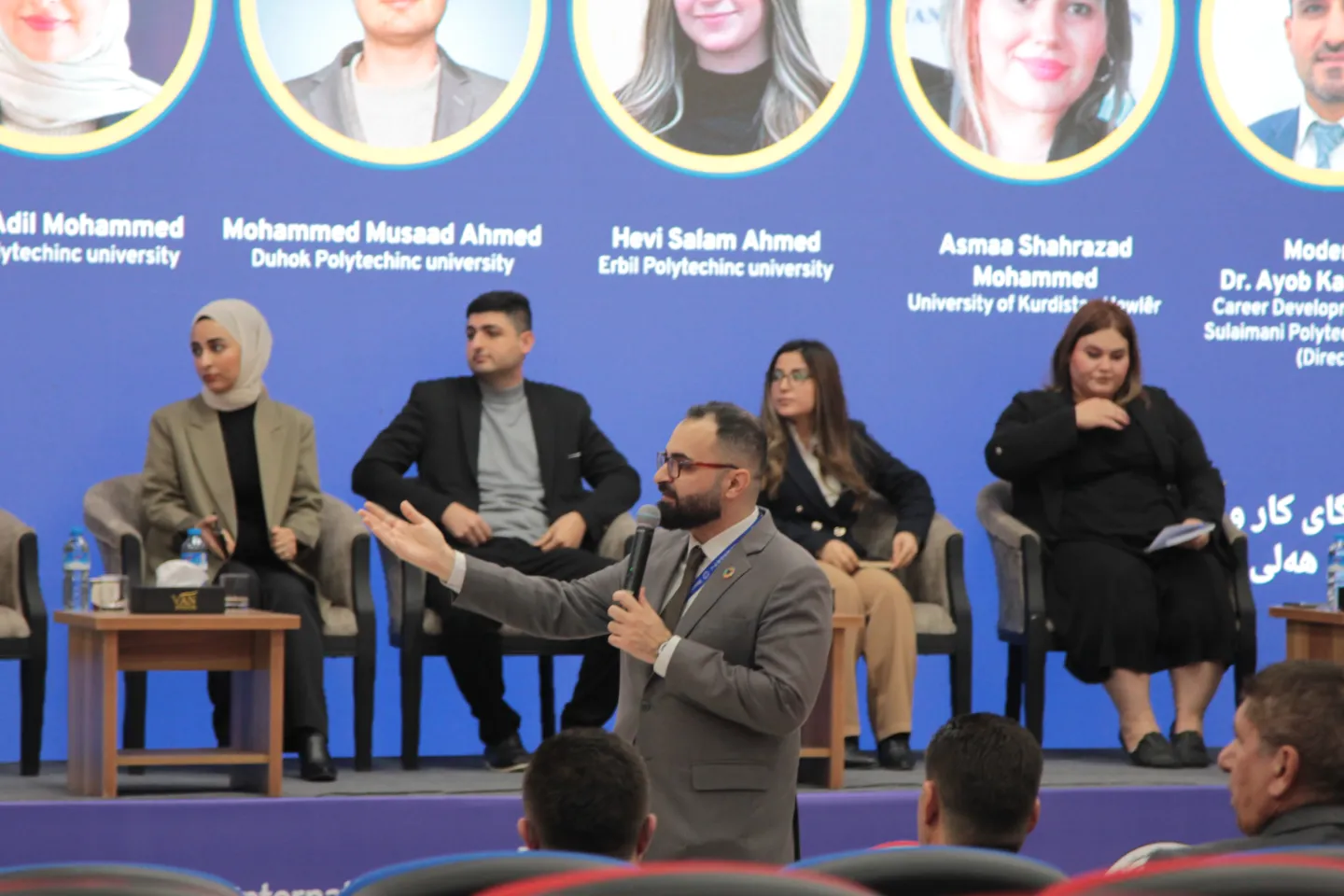 a man standing in front of a stage where other participants are seated for a panel discussion 