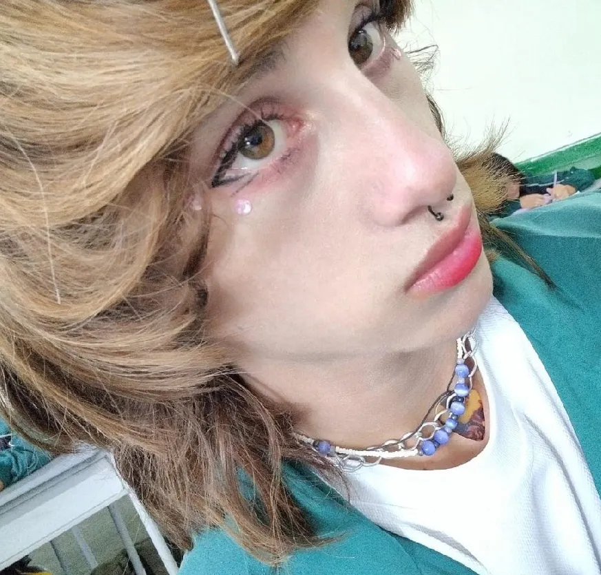 Joven sentado en el aula de clases luce un maquillaje suave. Al fondo otro joven concentrado en su actividad académica.