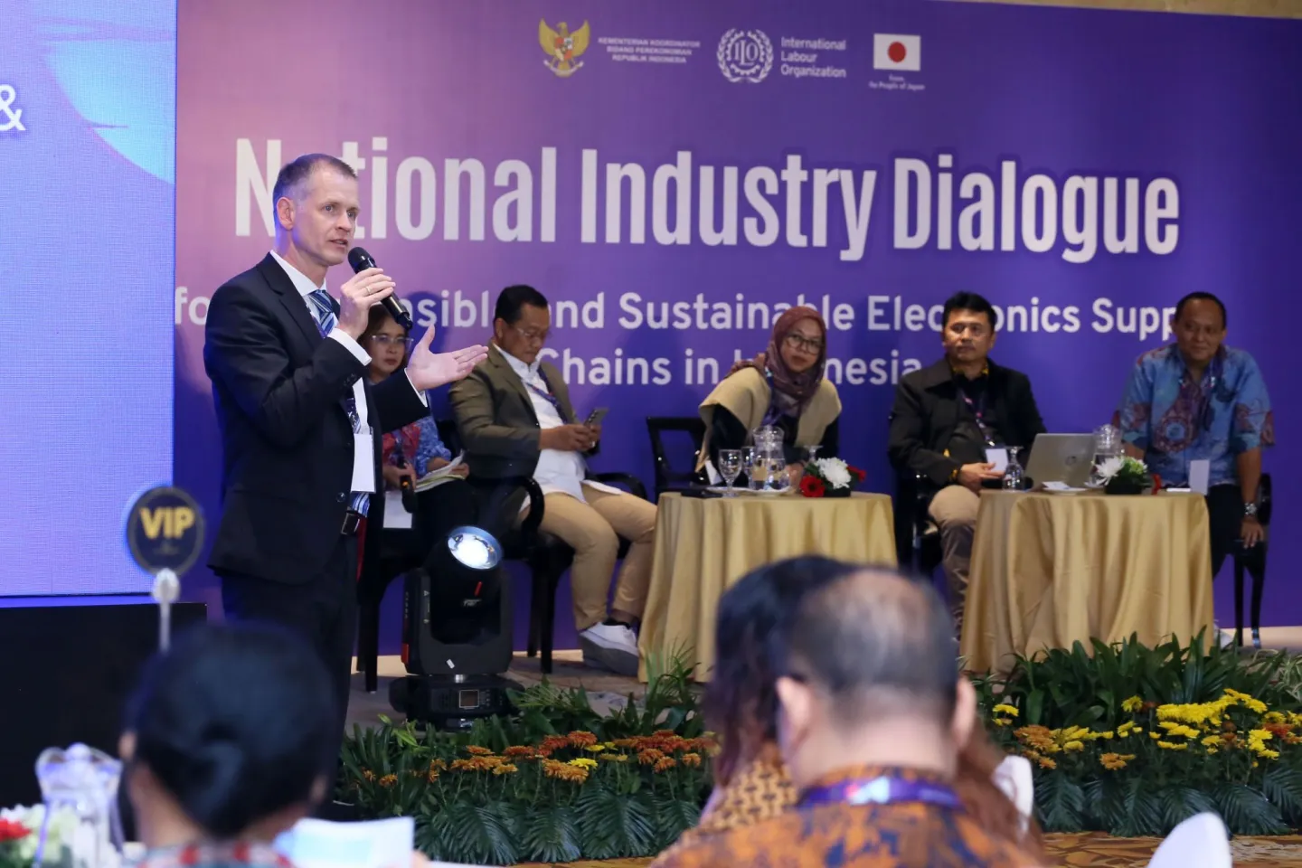Four people sitting on the stages with one of the speaker standing doing a presentation.