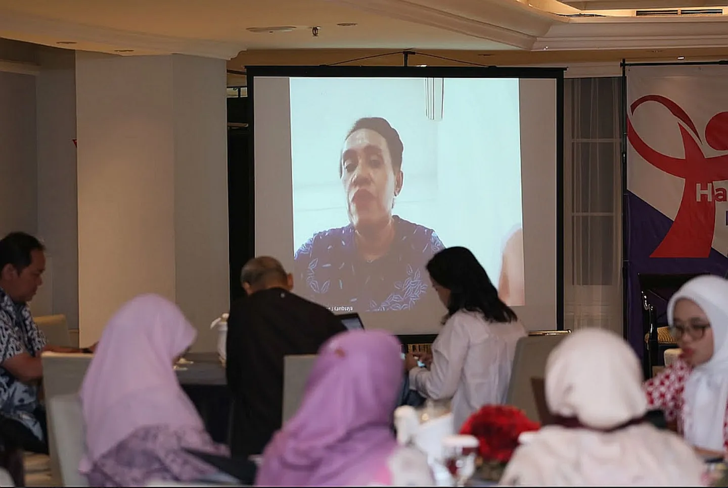 People attending meeting with screen highlighting speaker from outside Jakarta